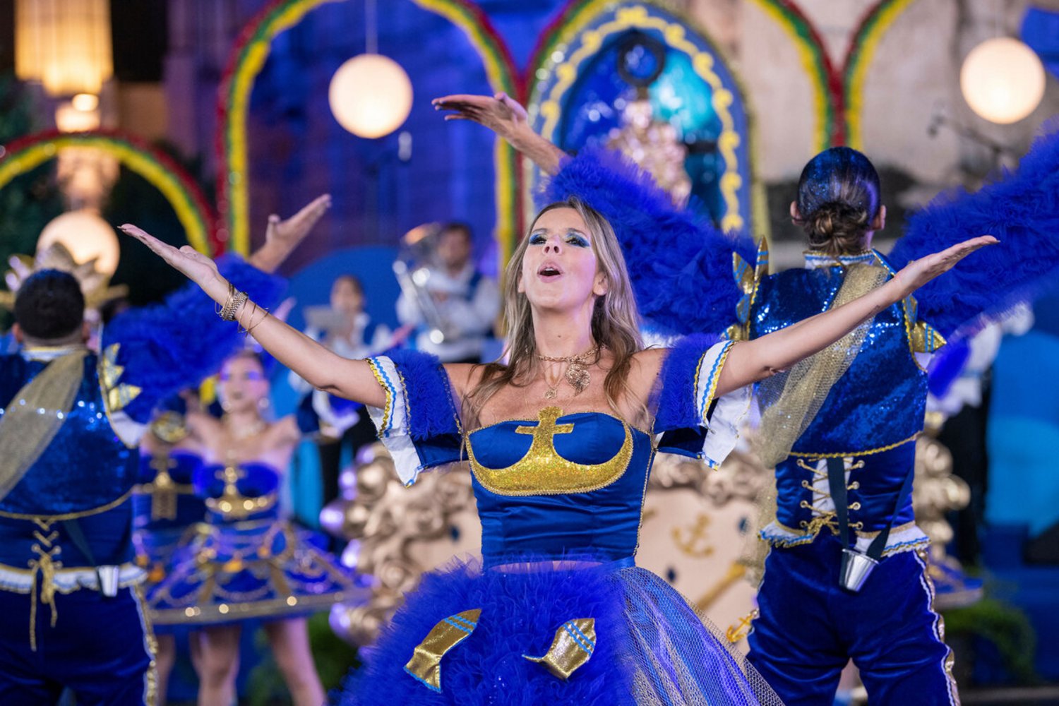 Mulher com vestido azul e dourado, braços abertos, a cantar e dançar no meio de um grupo de dançarinos num ambiente festivo e iluminado.